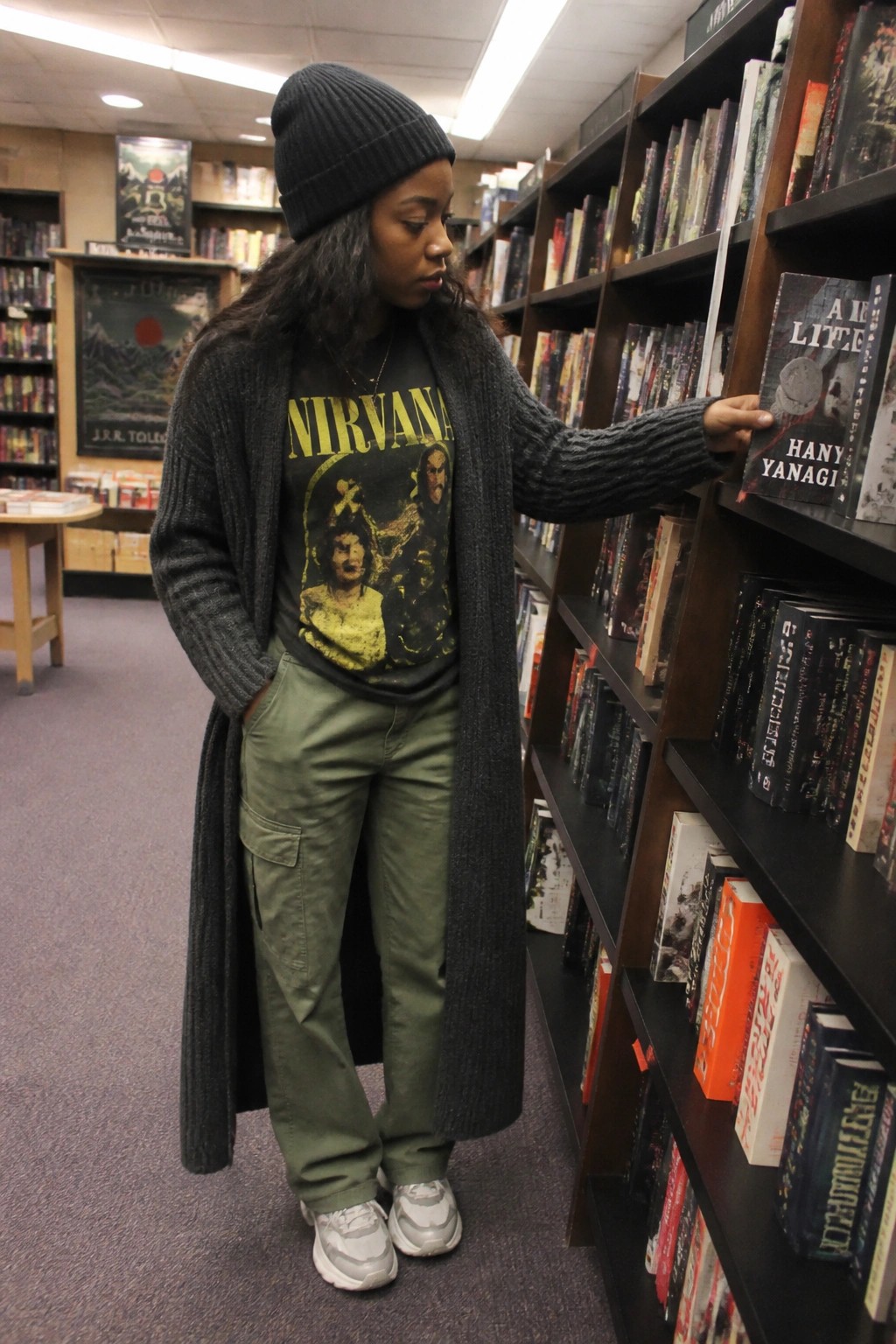 Woman in black beanie and Nirvana shirt reaches for book in library.