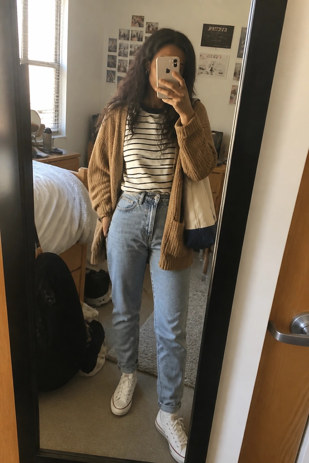 Woman in striped shirt and brown cardigan taking mirror selfie in jeans and white sneakers