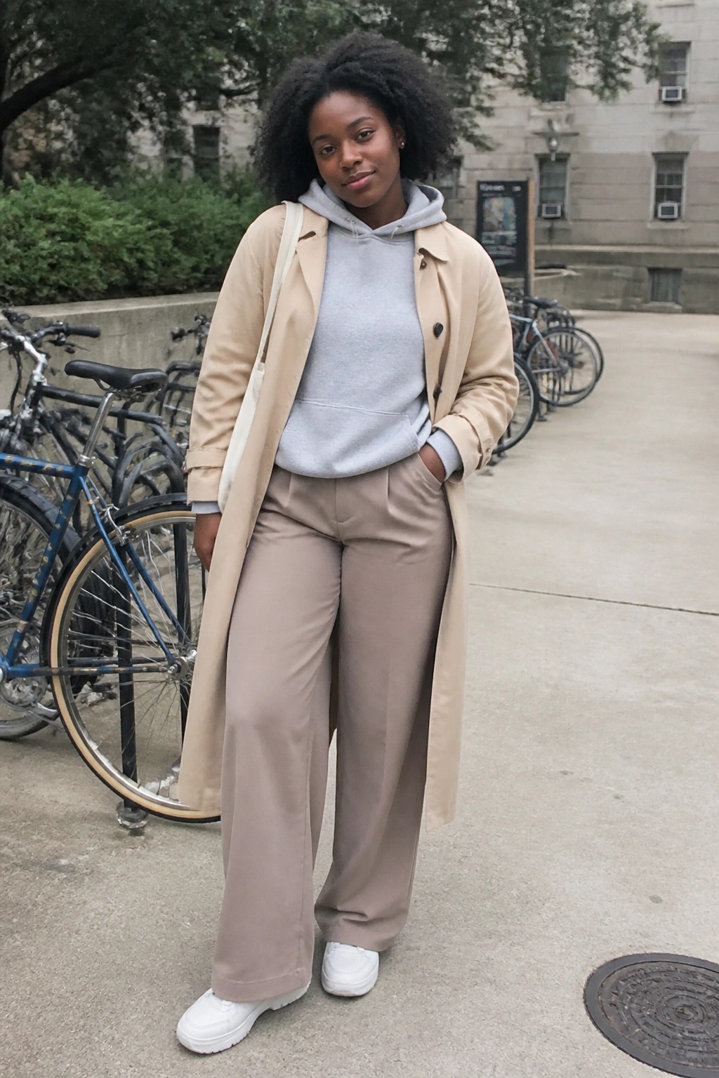 Young Black woman in beige trench coat over gray hoodie and wide pants outdoors.