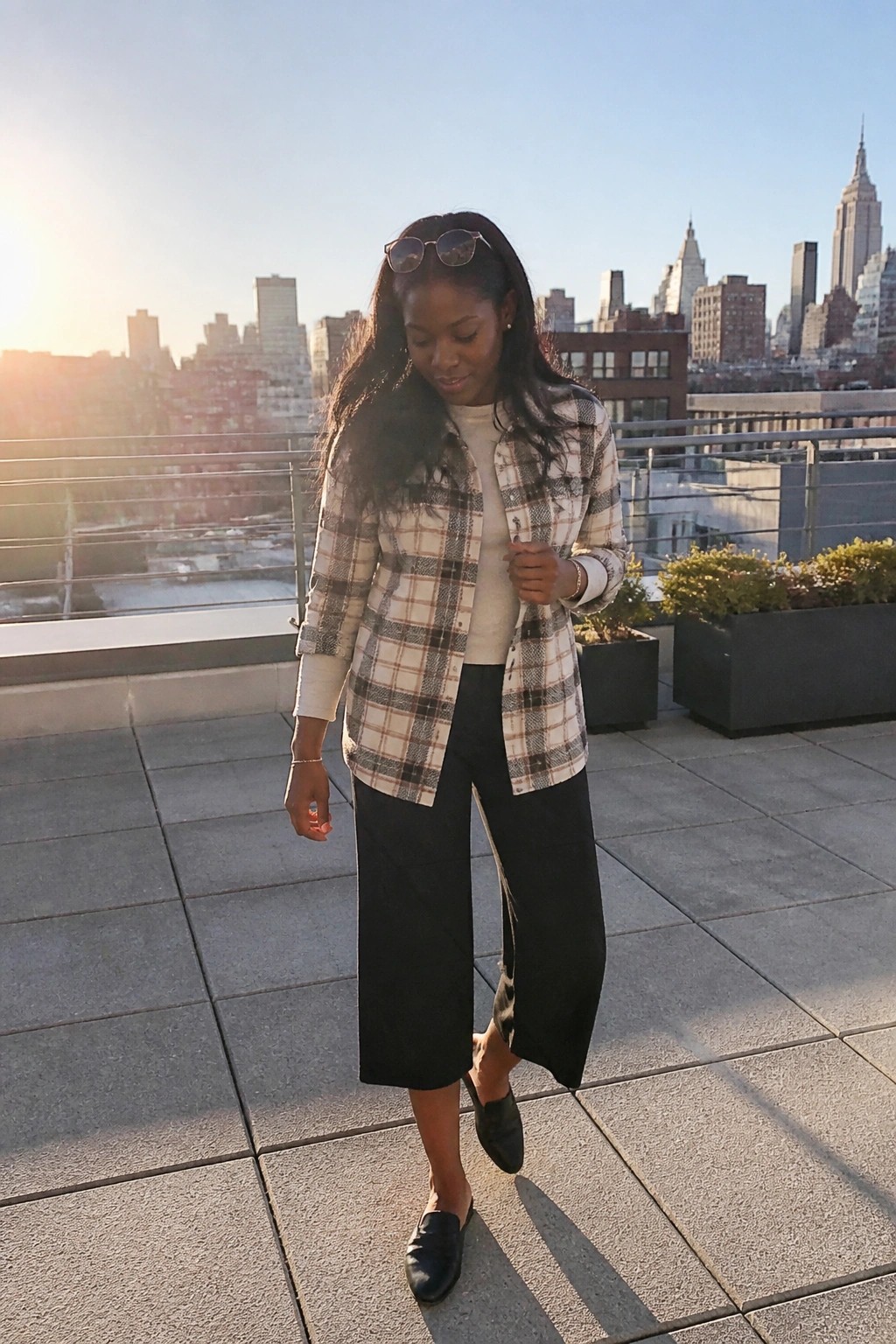 Woman in plaid jacket and black culottes on rooftop with city skyline.