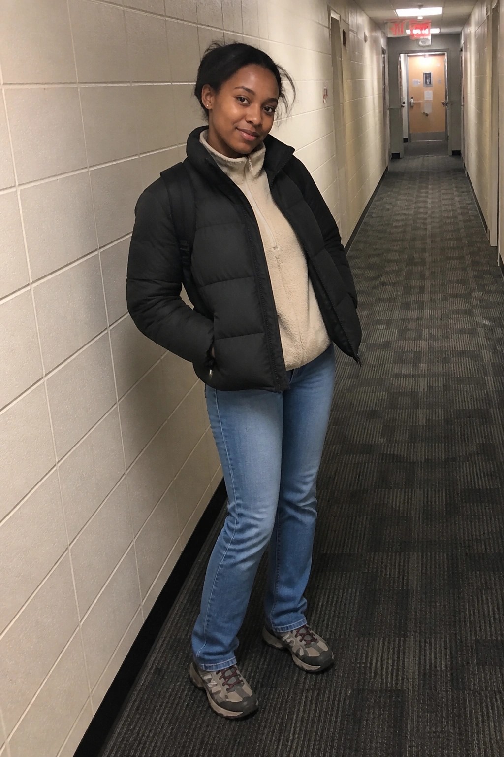 Young woman in black puffer vest and jeans leaning in hallway with backpack