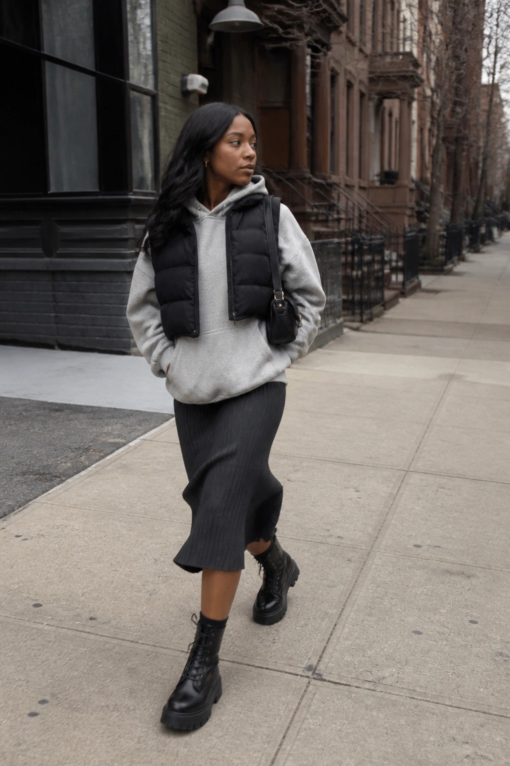 Woman in gray hoodie, black puffer vest, ribbed skirt, and combat boots on sidewalk