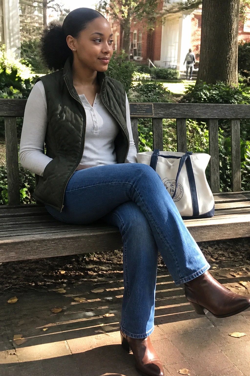 Woman in green quilted vest and blue jeans sits on bench with canvas tote bag