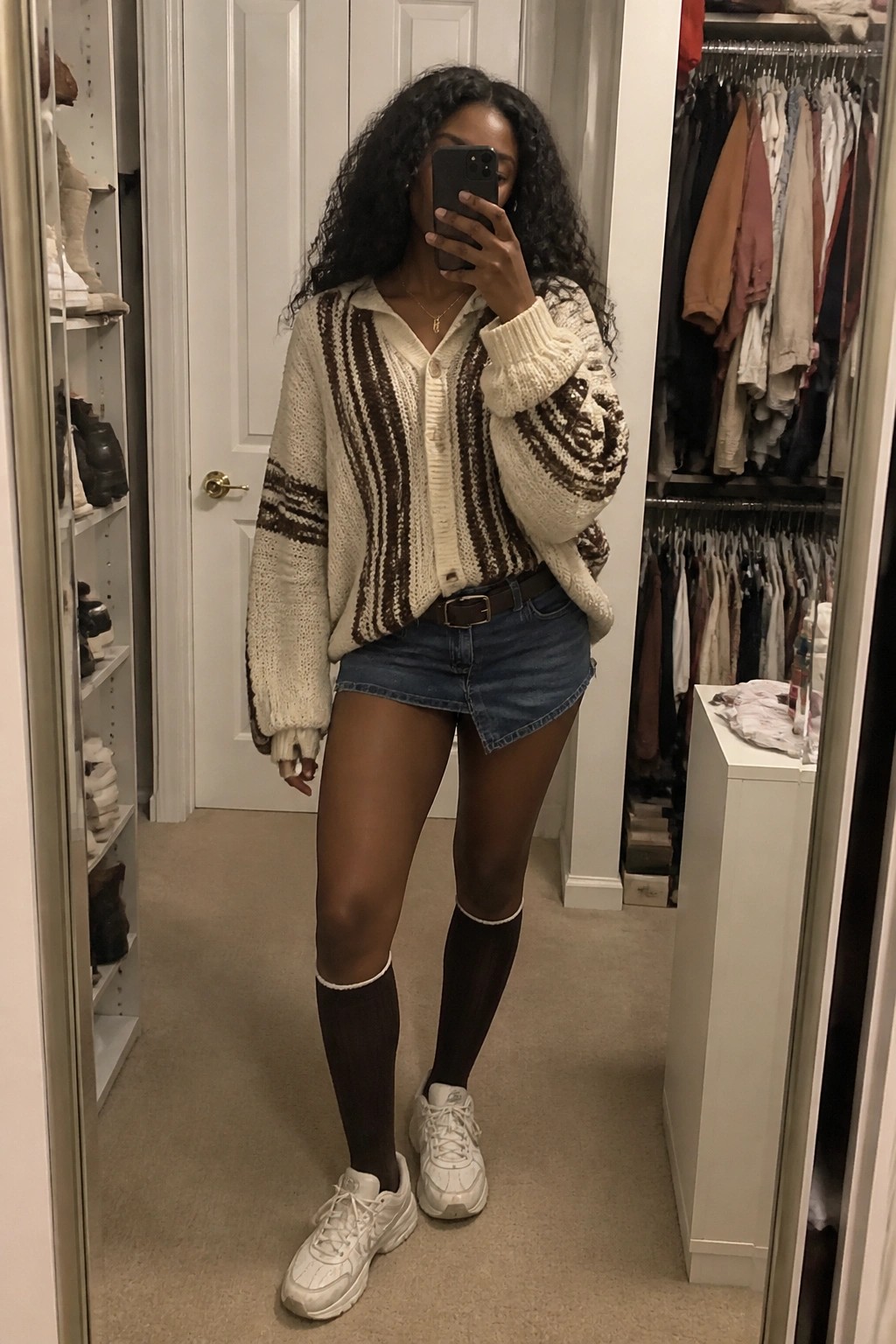 Woman in striped knit cardigan, denim skirt, and knee-high socks taking mirror selfie