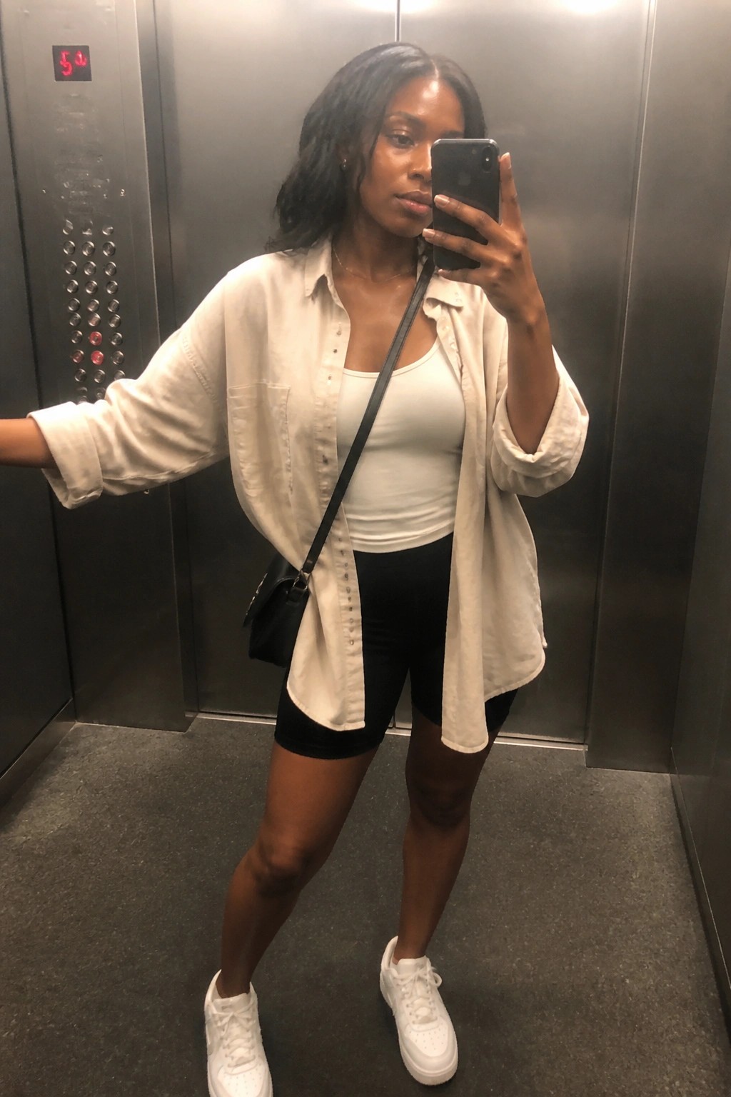 Woman in an elevator wearing an open oversized white button down shirt over a white tank top and black biker shorts, paired with white sneakers and a black crossbody bag.