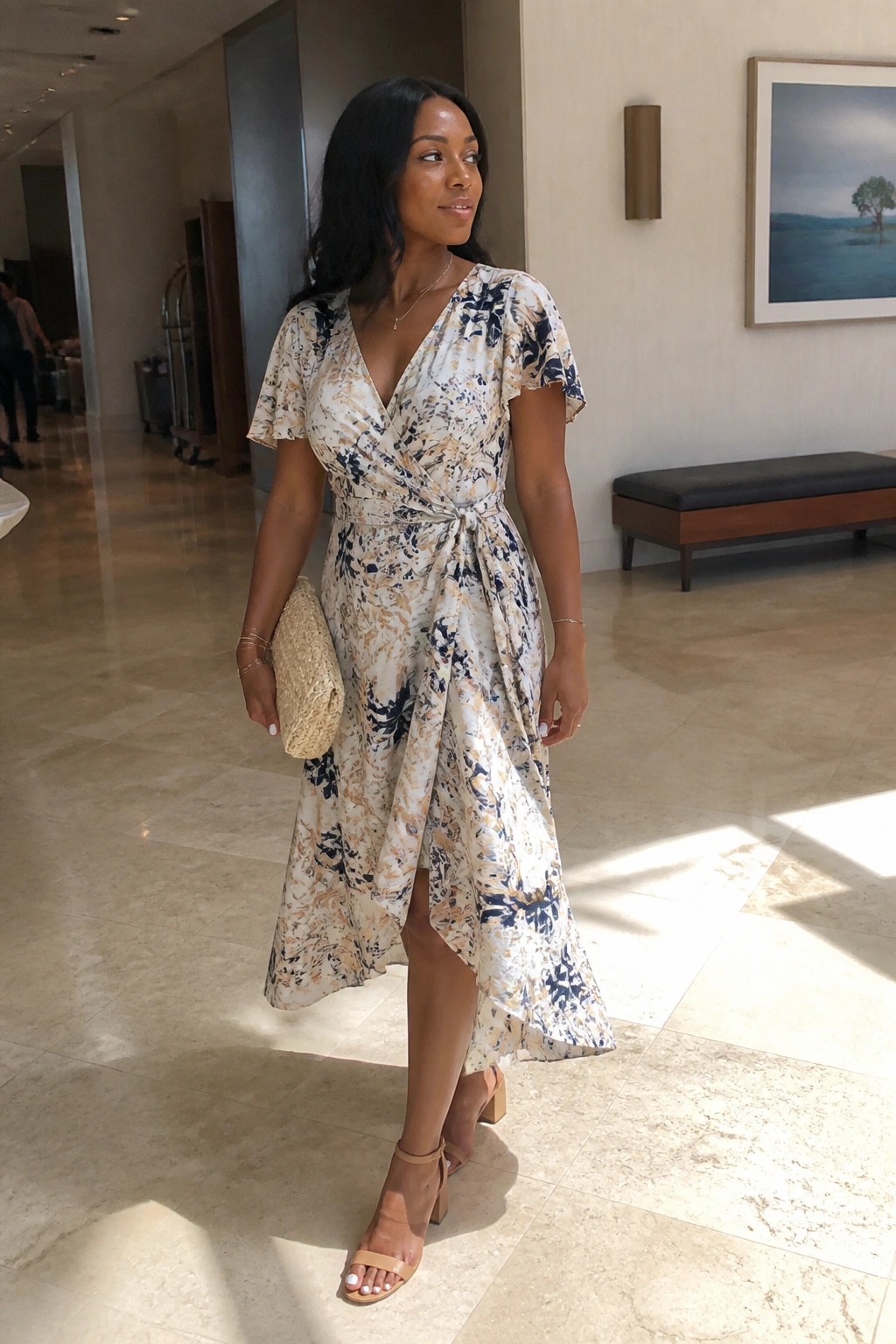Woman in floral wrap dress holding woven clutch in elegant lobby