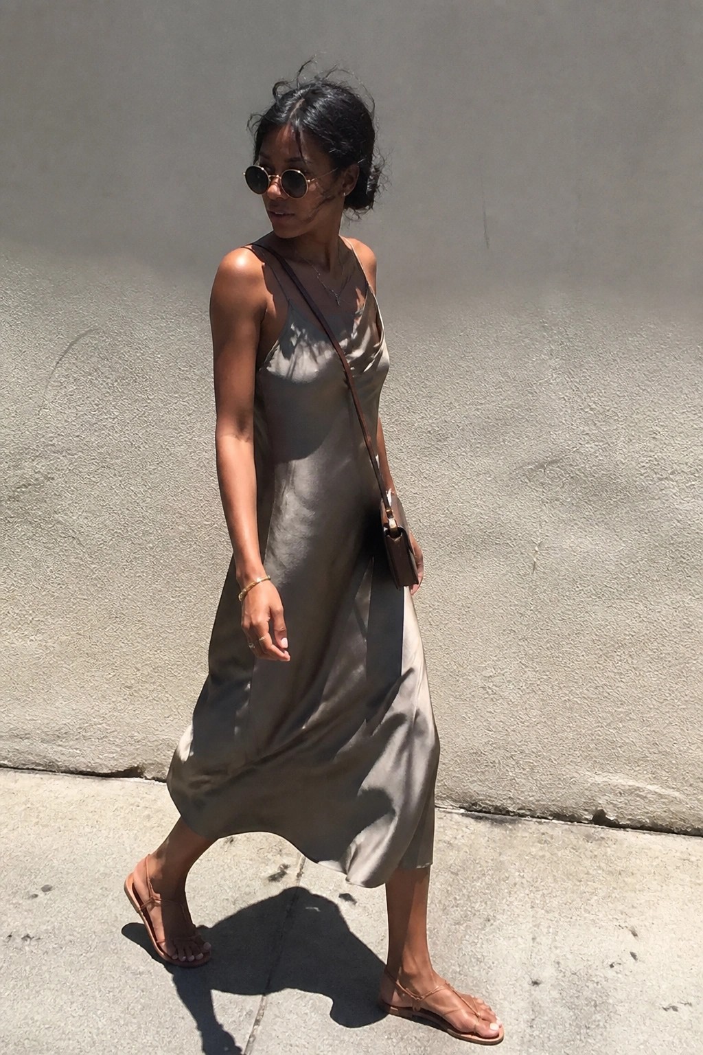Woman in flowing taupe satin slip dress and sandals walks on sunny sidewalk.