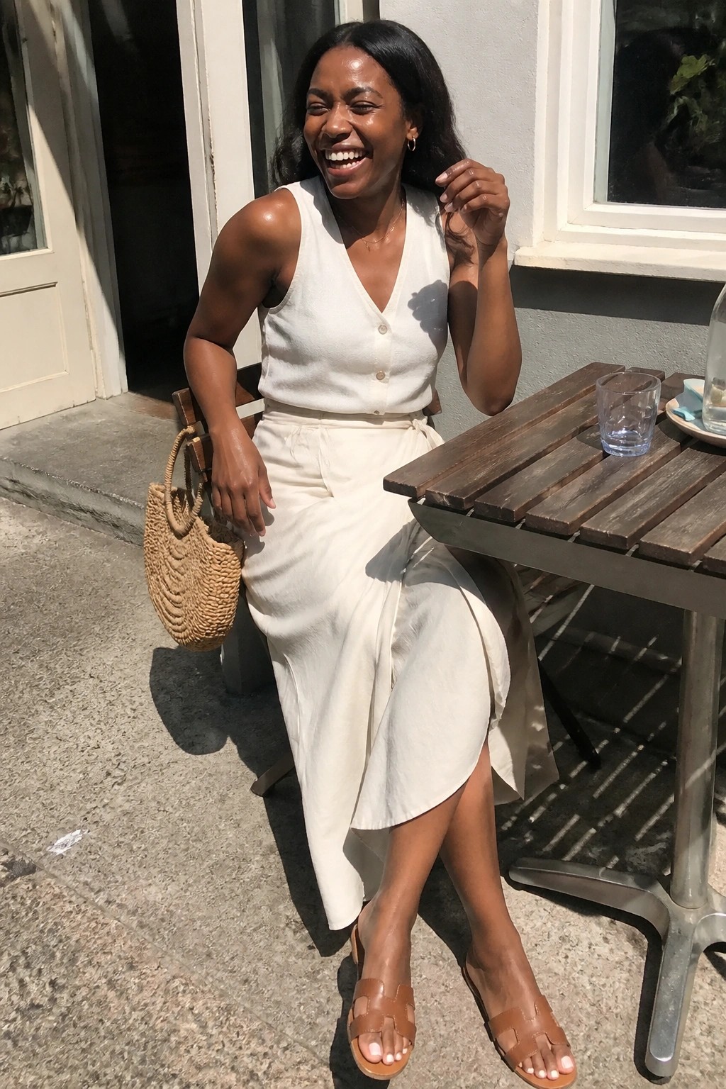 Smiling woman in white outfit at sunny outdoor table holding woven straw bag