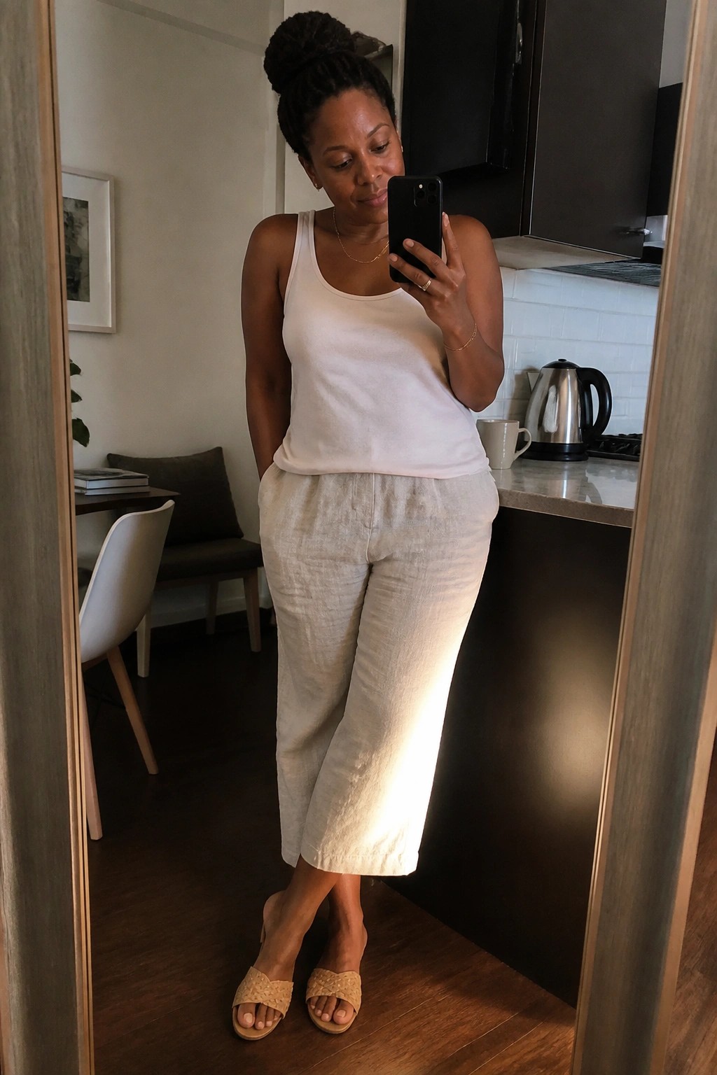 Woman wearing a white tank top with beige linen cropped pants and woven slides while holding a phone for a mirror selfie in a kitchen setting.