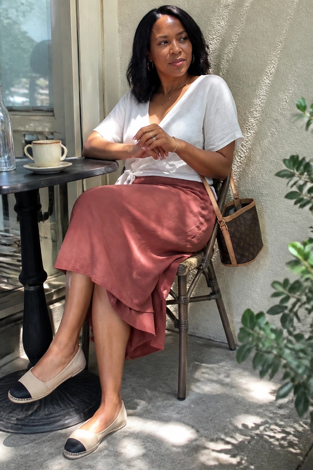 Woman wearing a white short sleeve wrap top tucked into a dusty rose midi skirt with asymmetrical hem, paired with beige and black espadrille flats and carrying a patterned handbag.