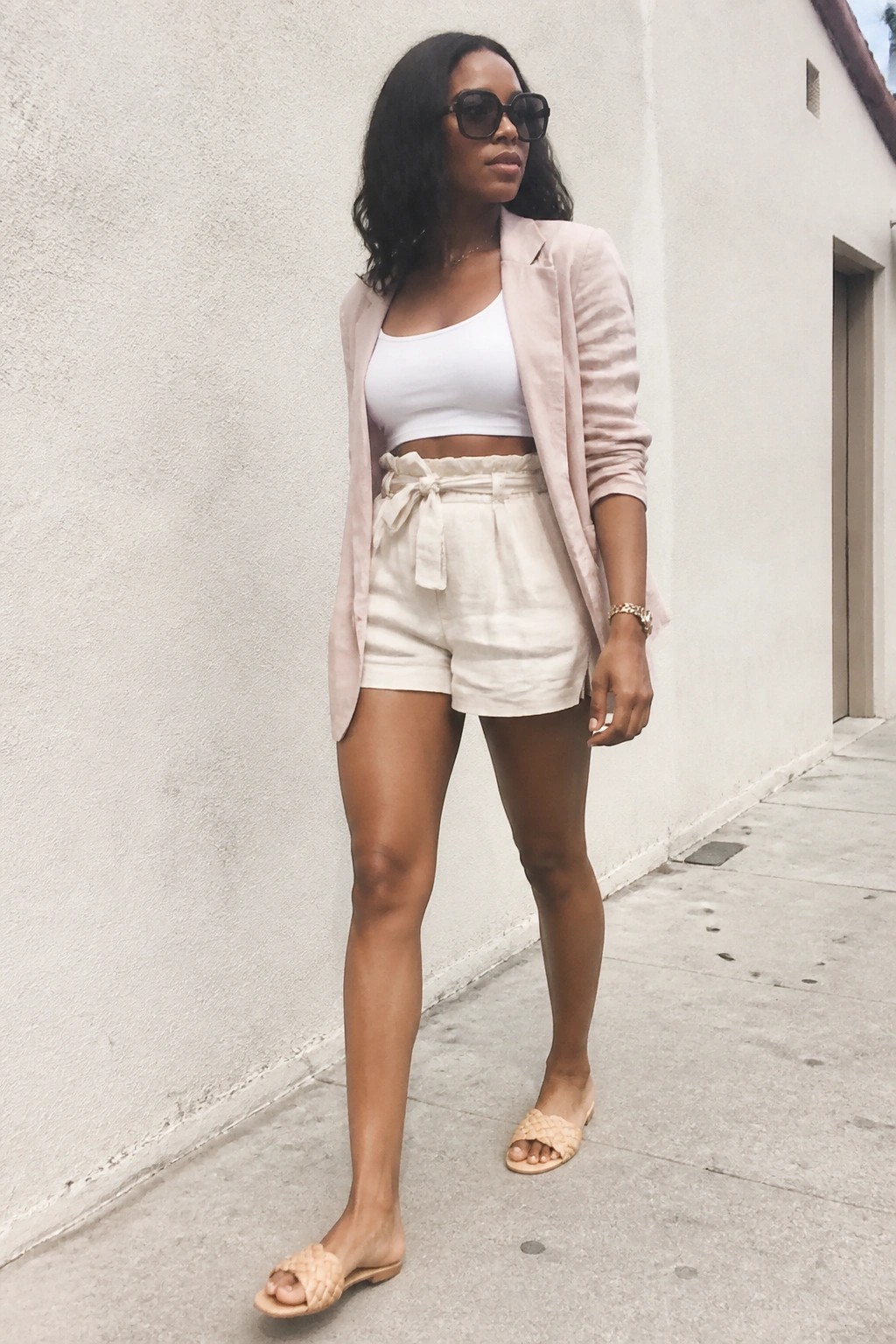 Woman in pink blazer, white crop top, and white shorts with woven sandals
