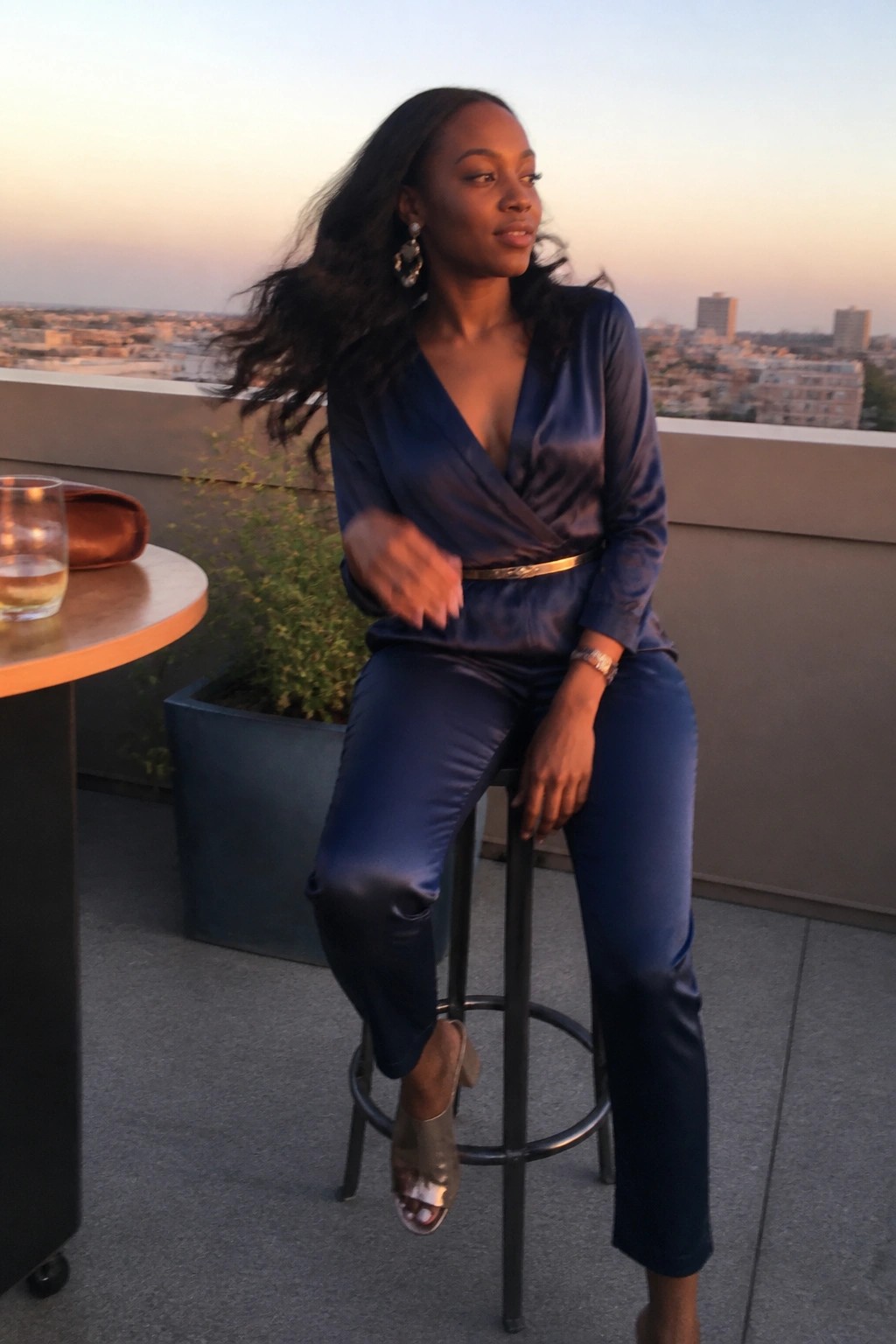 Woman in blue satin jumpsuit on bar stool at sunset rooftop with city view.