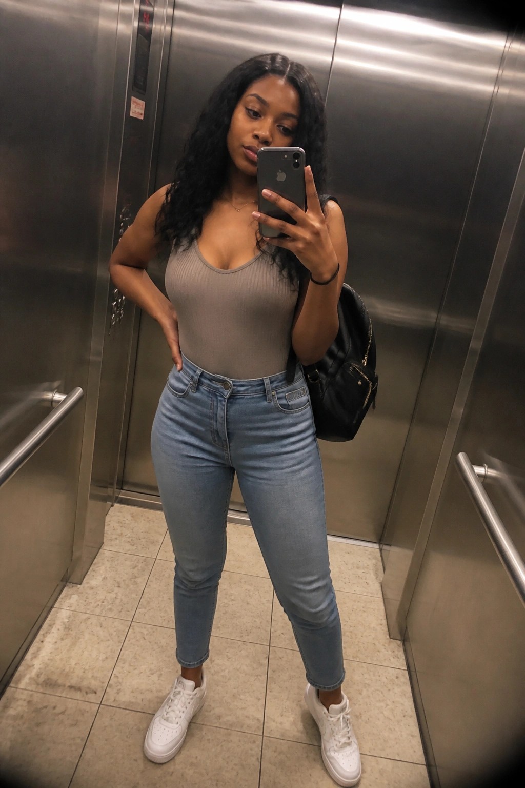 Woman with long dark hair in gray bodysuit and blue jeans taking elevator selfie.