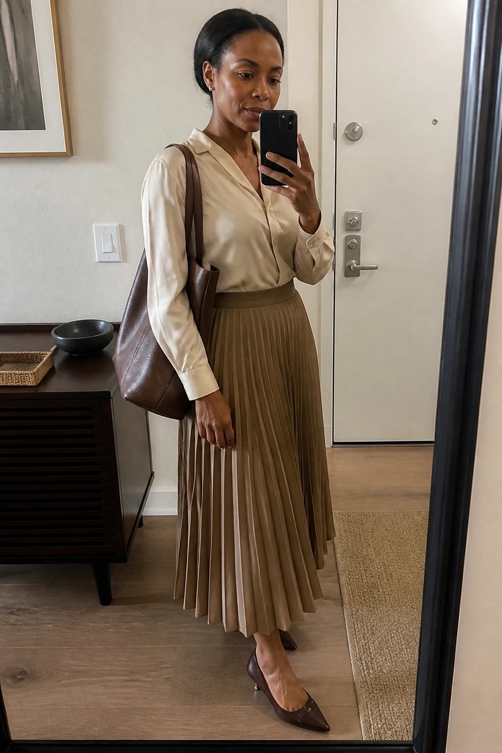 Woman in beige blouse and pleated skirt taking mirror selfie with brown bag