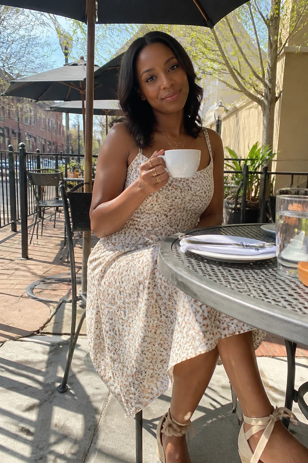 Woman wearing a cream floral midi dress with thin straps, gold necklaces and rings, and beige strappy block heels while seated at an outdoor table holding a white cup.