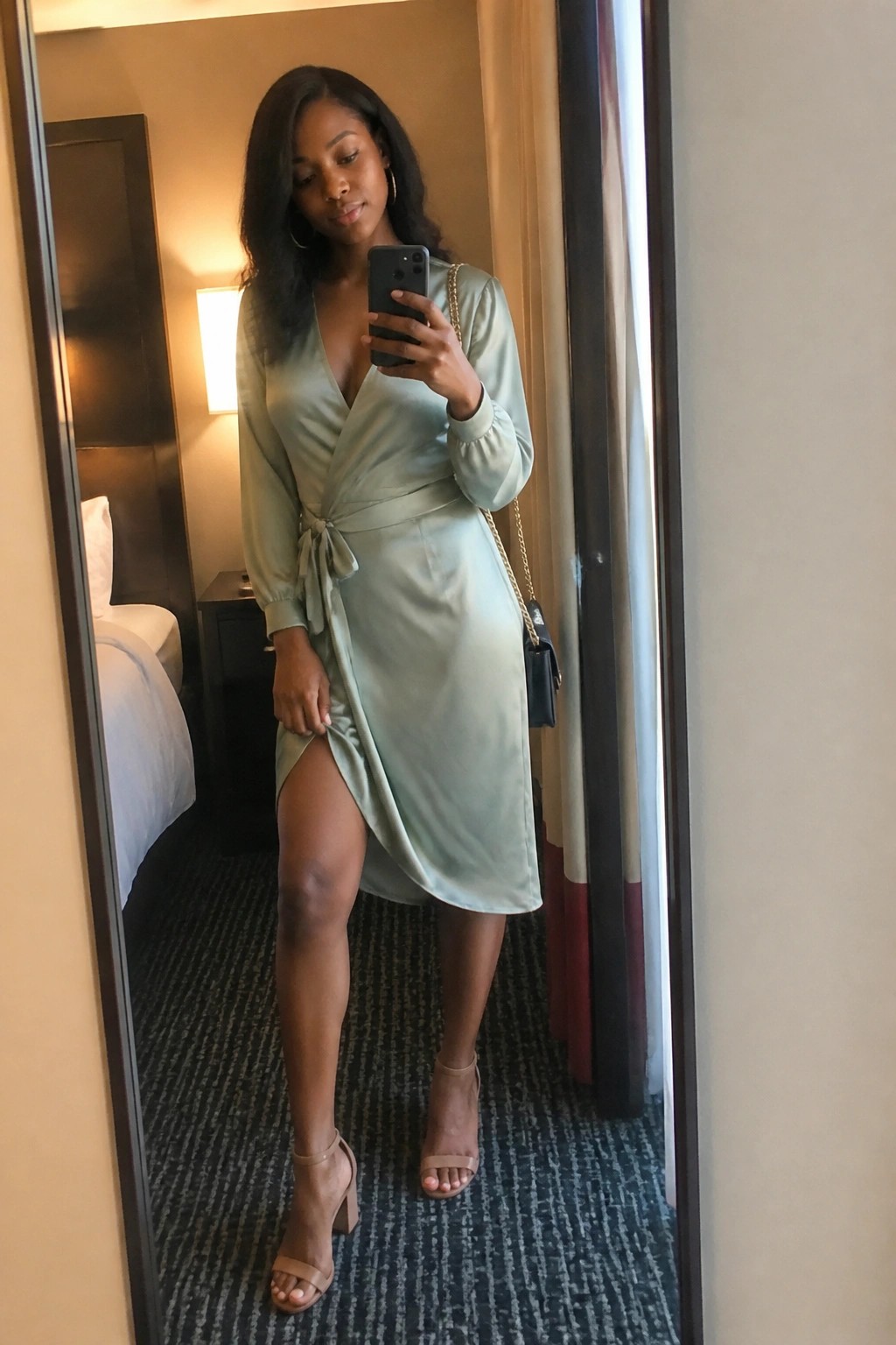 Woman in sage green satin wrap dress taking mirror selfie in hotel room