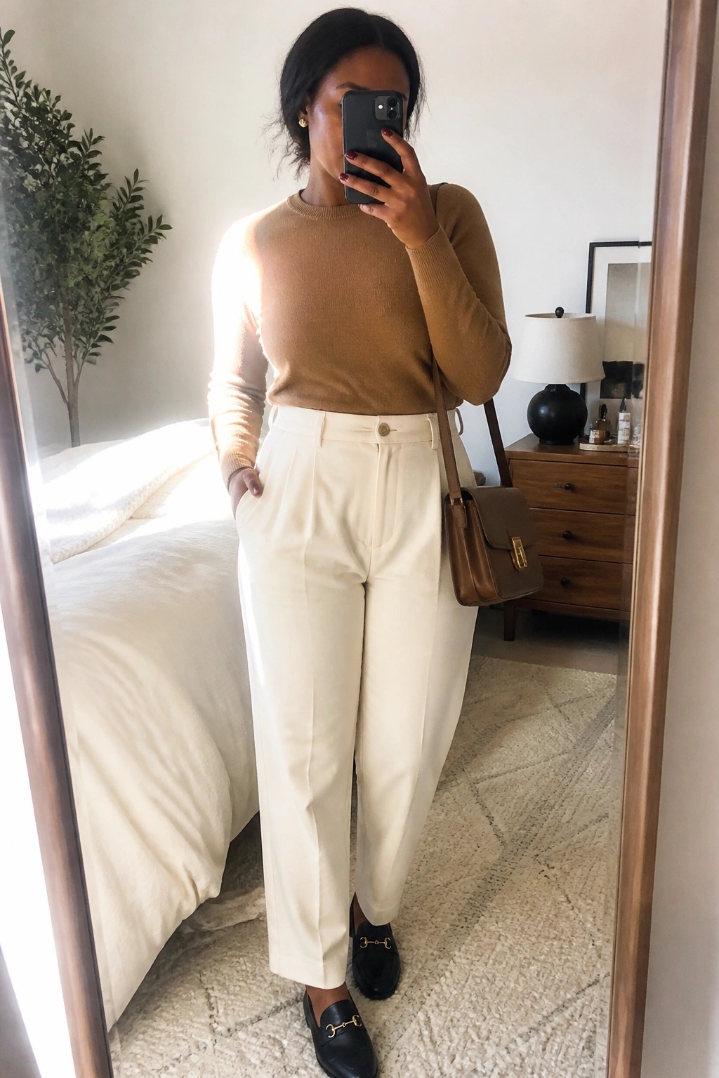 Woman in tan sweater and white trousers taking mirror selfie in bedroom.