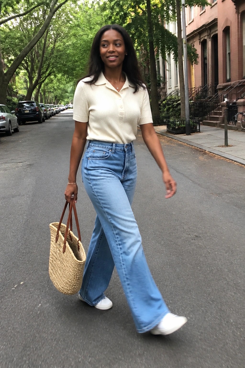 Woman wearing a cream short sleeve polo knit sweater tucked into light blue wide leg jeans with white sneakers and carrying a woven straw tote bag.