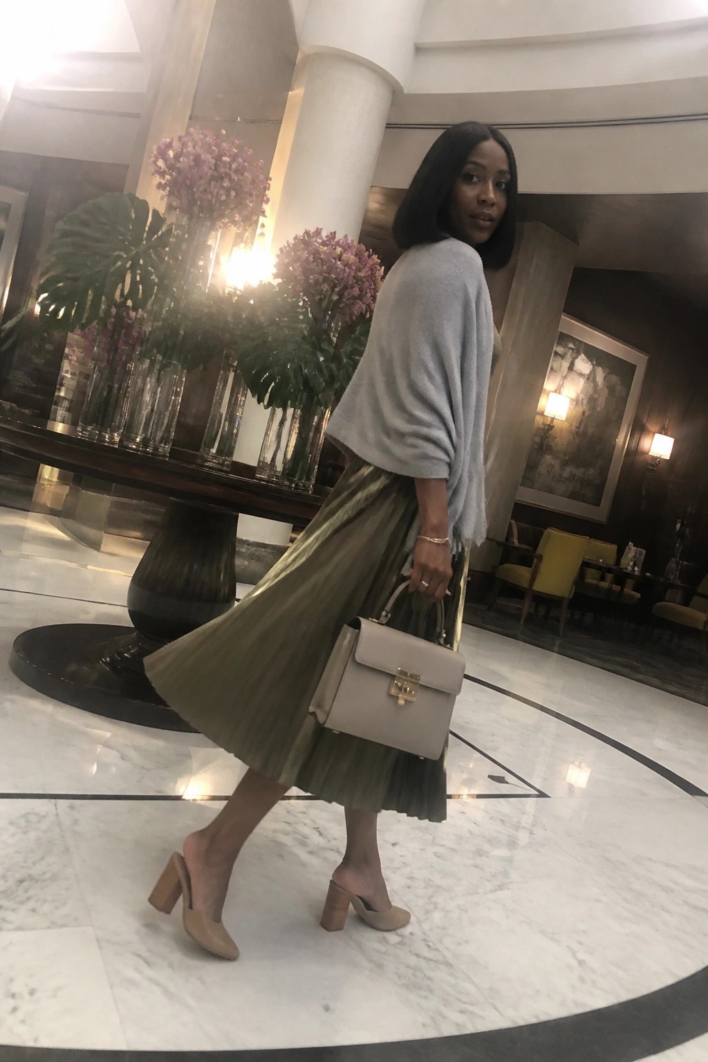 Woman in gray sweater and green pleated skirt holding beige handbag in marble lobby