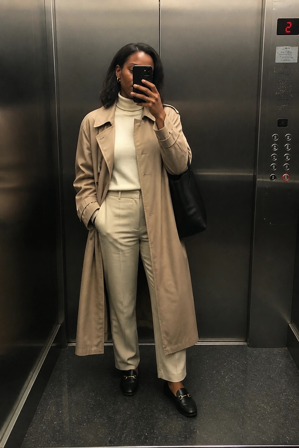 A Black woman wearing a beige trench coat over a cream turtleneck and matching wide-leg trousers with black loafers holding a black bag in an elevator selfie.