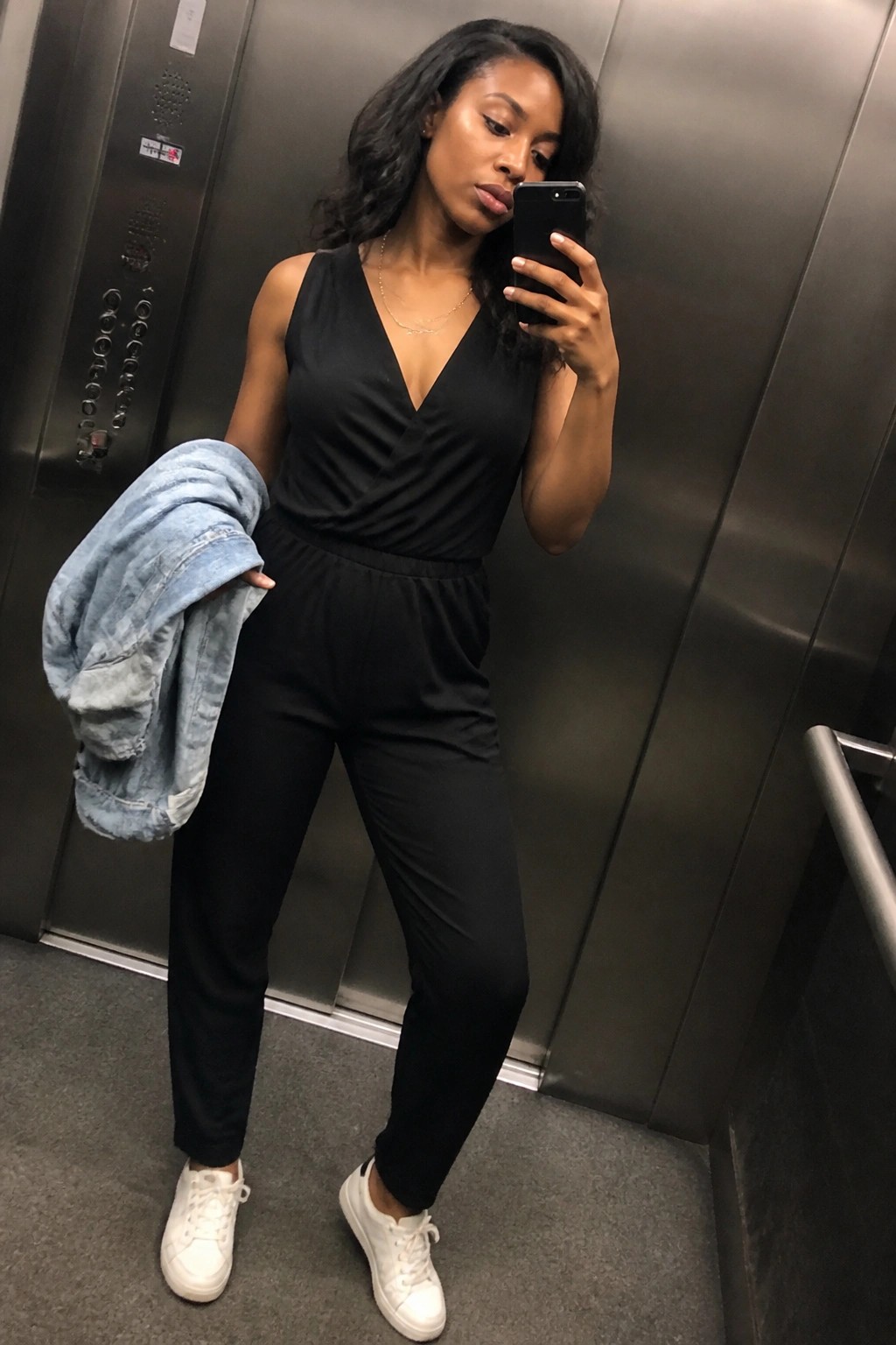 Woman in black jumpsuit takes selfie in elevator holding denim jacket