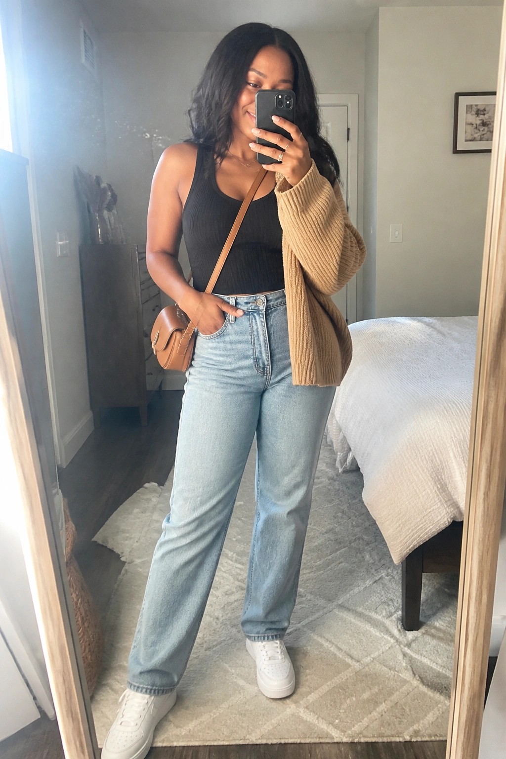 Woman in black tank top and tan cardigan taking bedroom mirror selfie