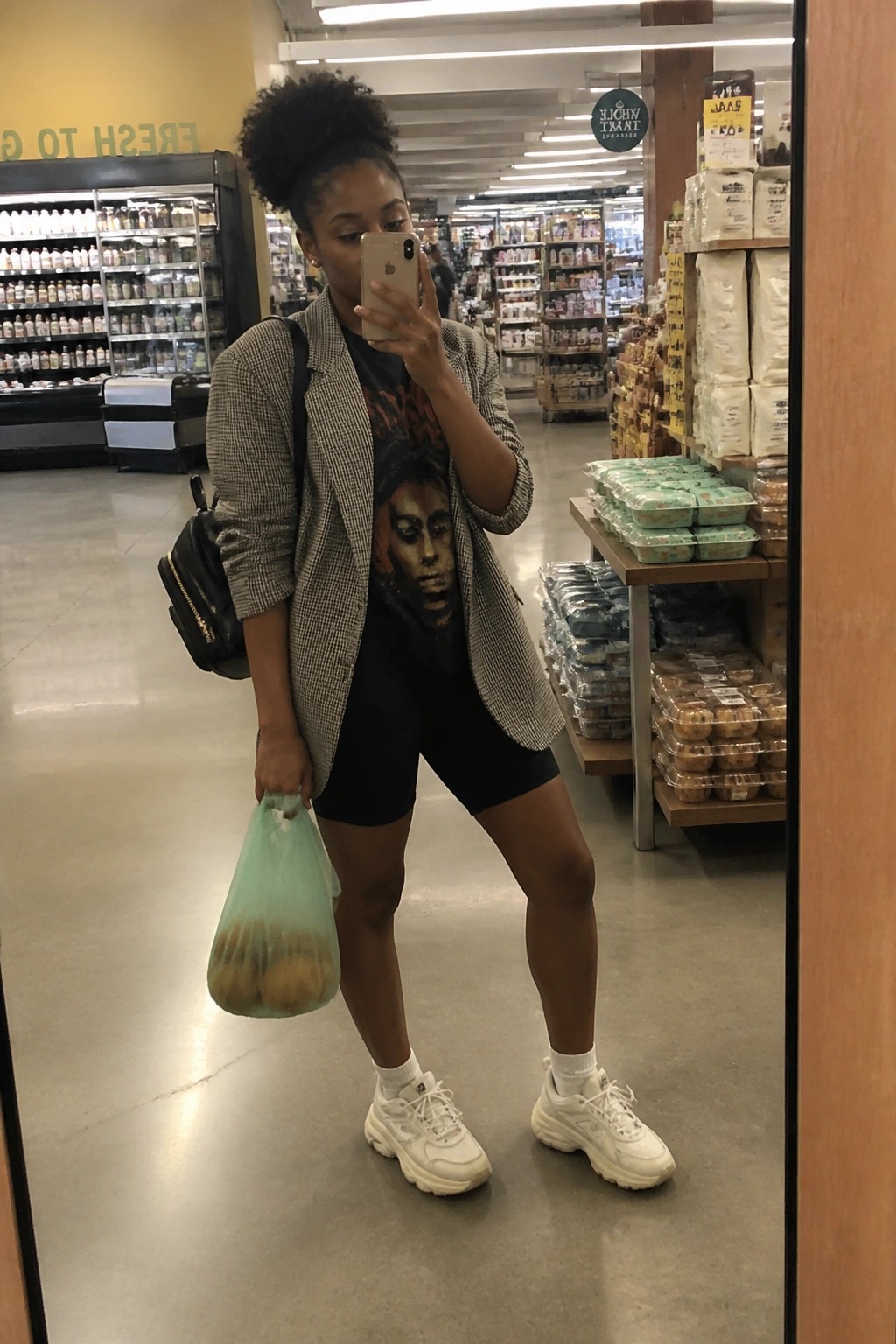Woman in checkered blazer and biker shorts takes mirror selfie holding oranges in store