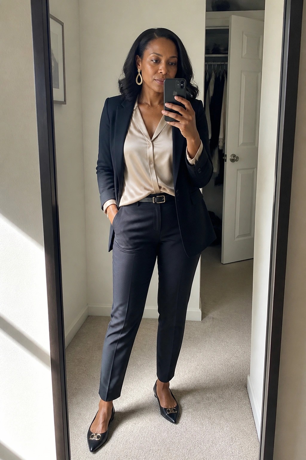 Woman in black pantsuit and beige blouse taking mirror selfie with iPhone.