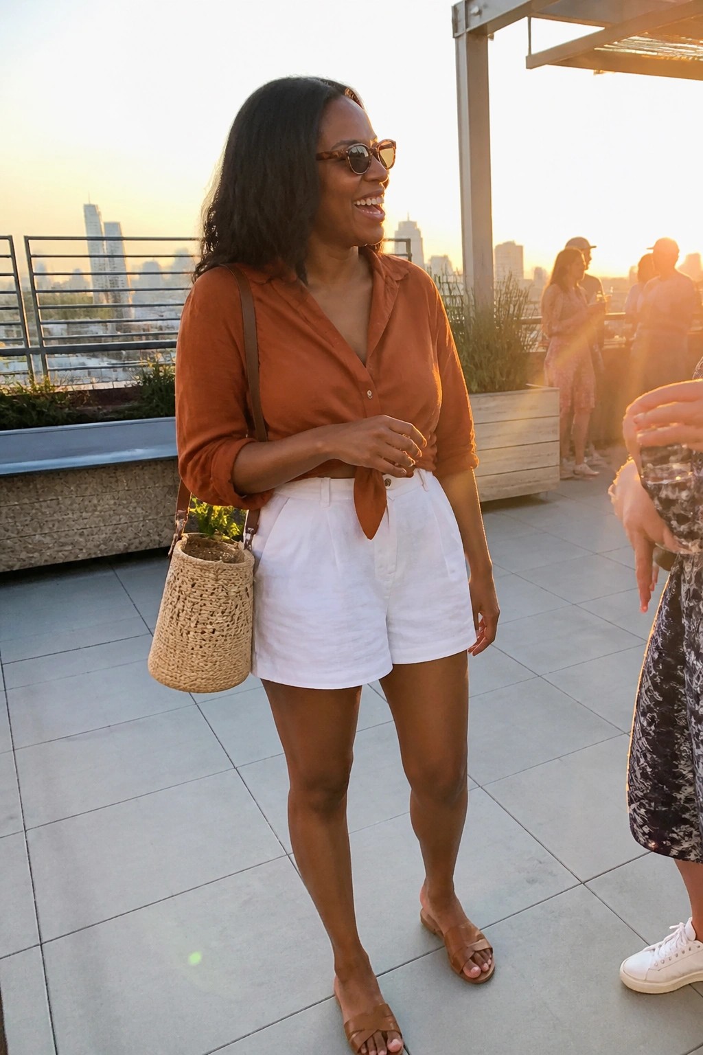 Woman in orange tied shirt and white shorts with woven bag on sunset rooftop