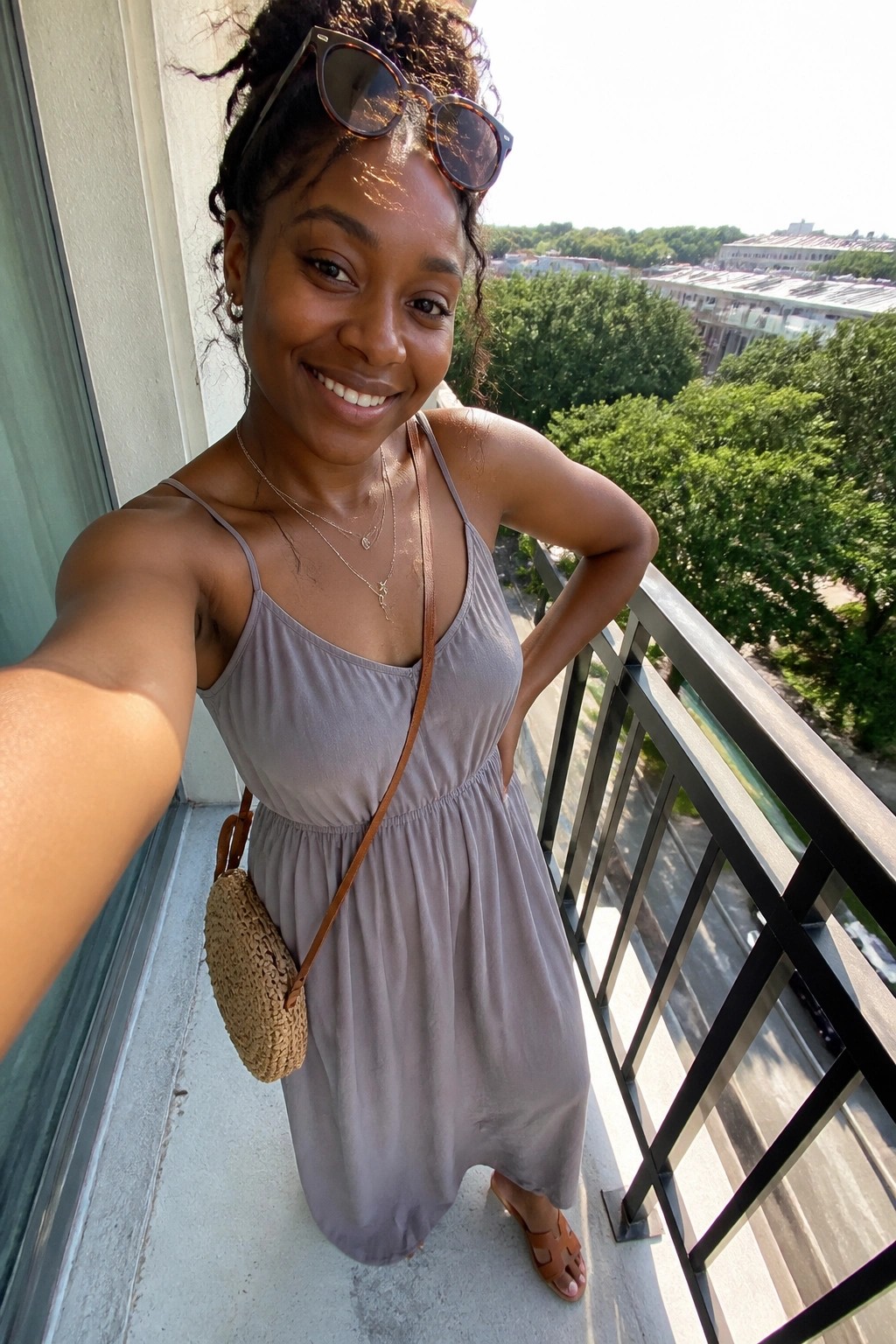 Smiling woman in light purple sundress on balcony with sunglasses and woven bag