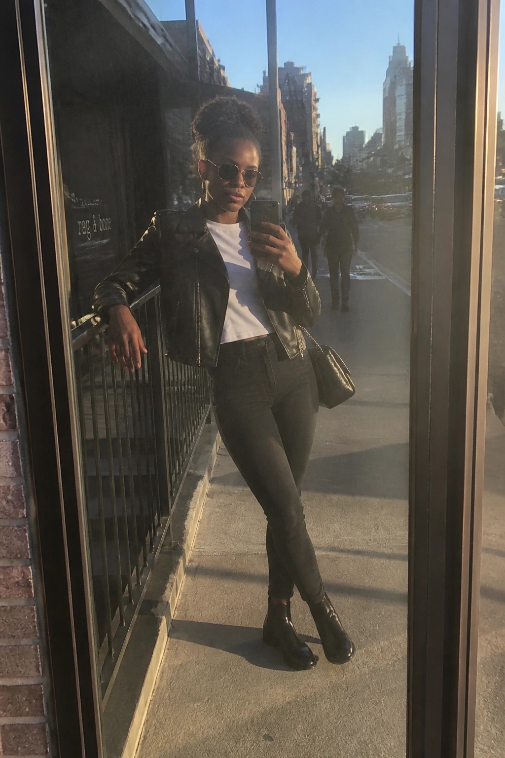 Woman in black leather jacket and sunglasses taking mirror selfie on city street