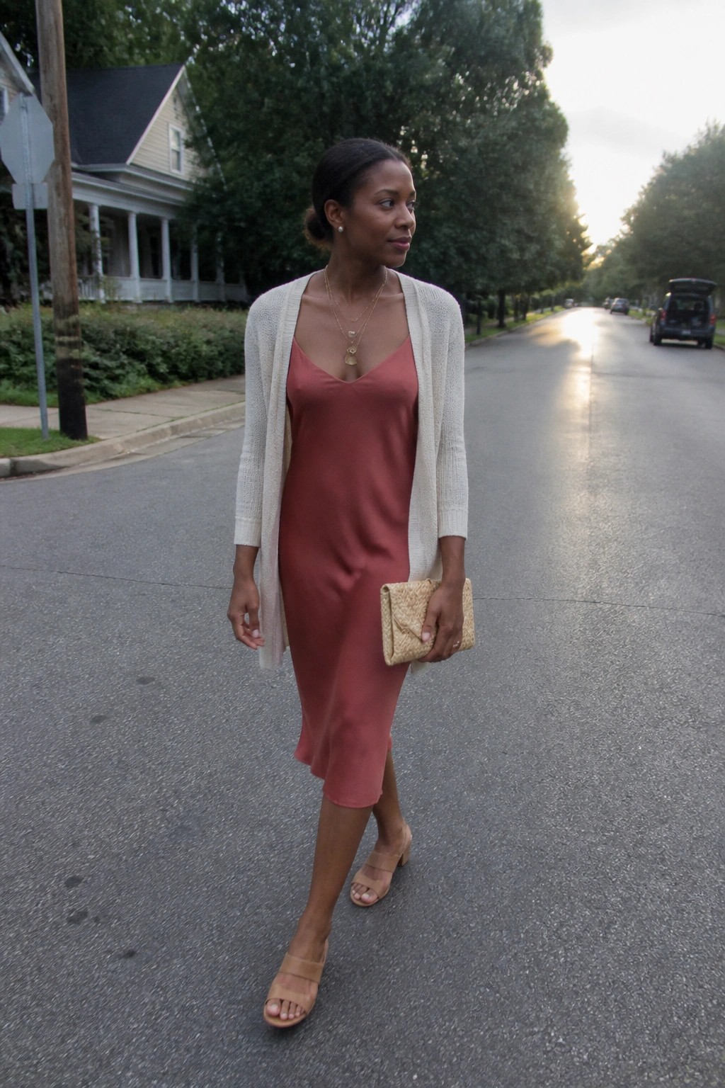 Woman in rose slip dress and cream cardigan holding woven clutch on suburban street