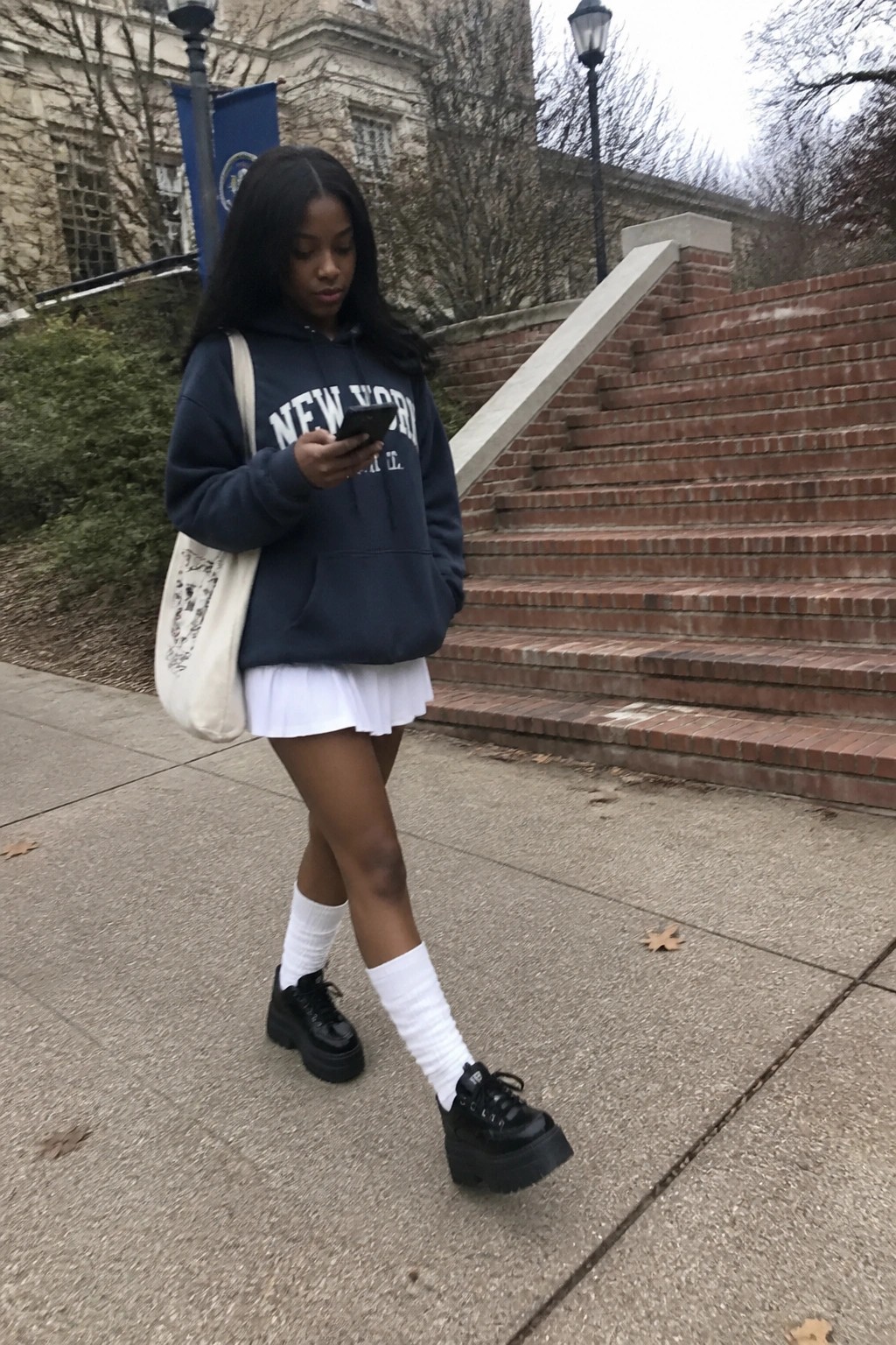 Woman in navy New York hoodie and white skirt views phone near brick steps