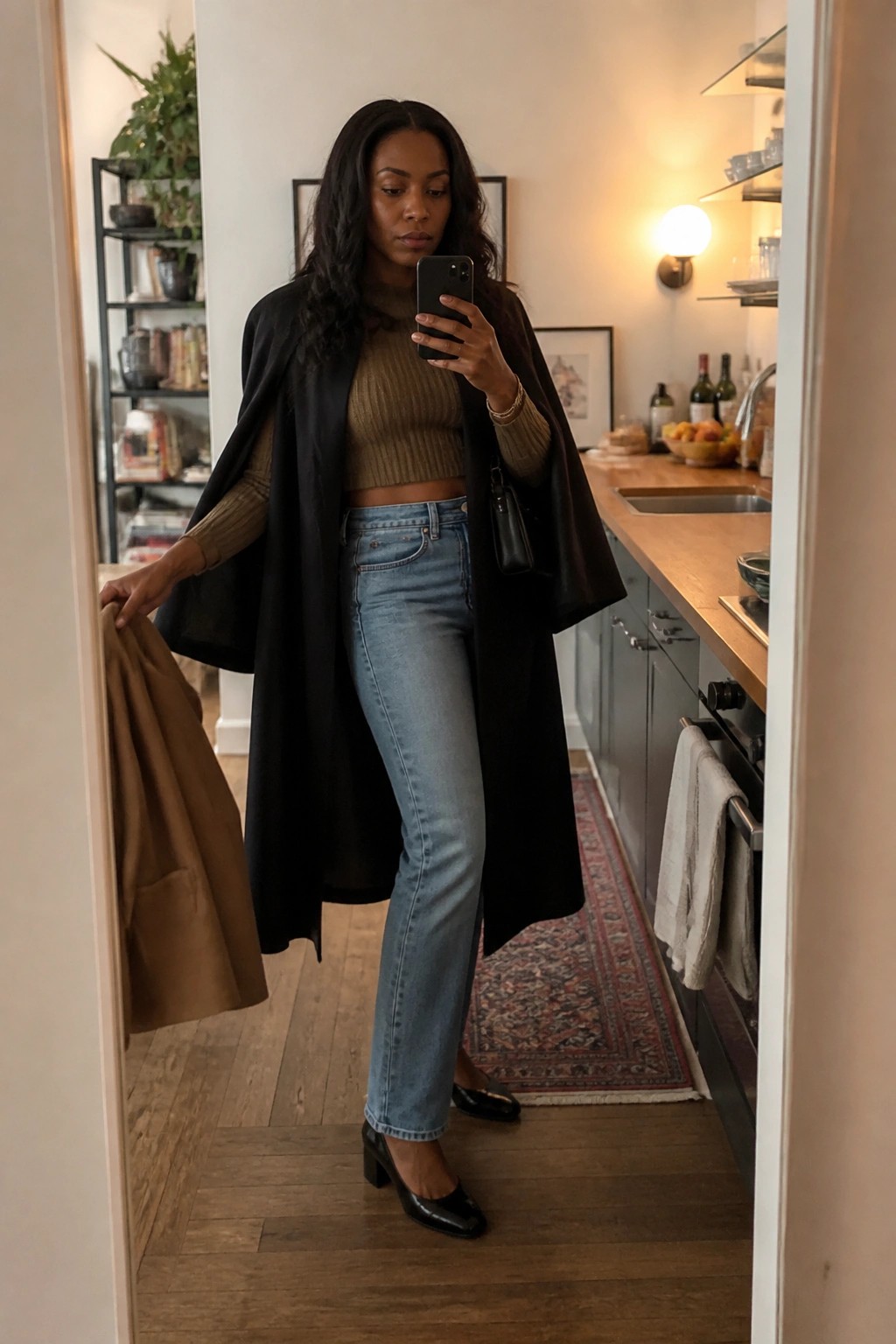 Woman in a long black cape layered over a taupe ribbed crop sweater and light blue straight leg jeans with black patent block heel shoes holding a phone for a mirror selfie.
