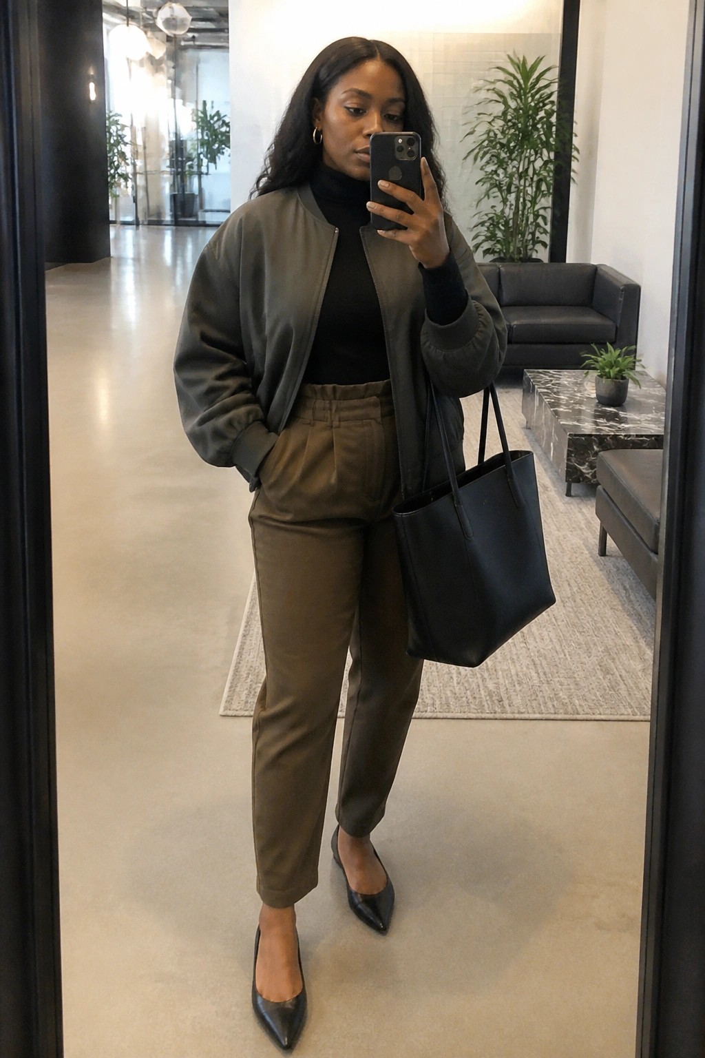 Woman in olive bomber jacket and pants taking mirror selfie with black tote bag