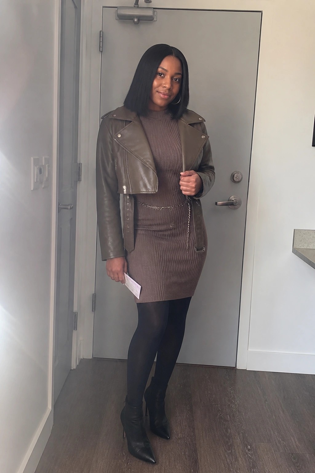 Woman in brown ribbed dress and olive leather jacket by gray door with black boots.