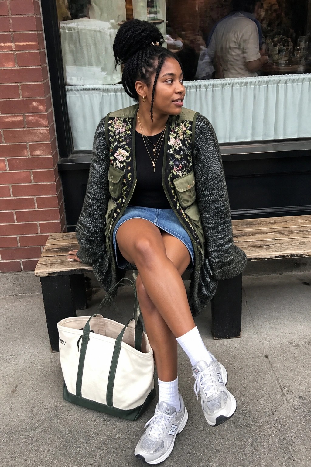 Woman in embroidered floral vest and denim skirt sits on bench with tote bag