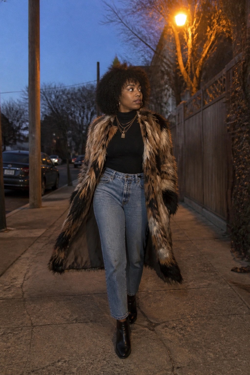 Woman in long fur coat and jeans walks on city sidewalk at night.