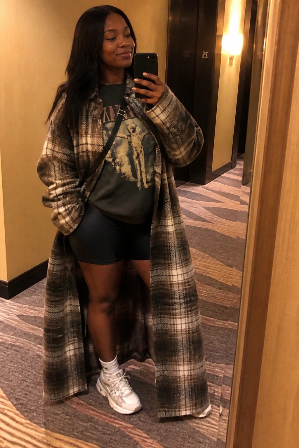 Woman with long dark hair in plaid coat and biker shorts taking mirror selfie