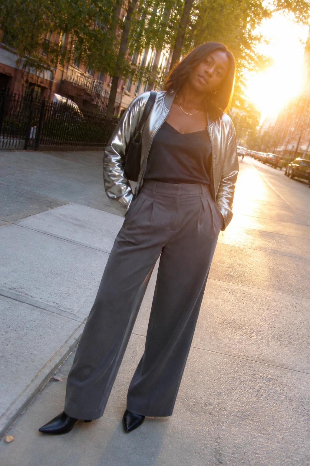 Woman in silver metallic jacket and gray wide-leg pants on city sidewalk at sunset