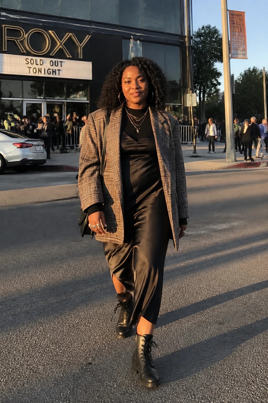 Woman in plaid blazer over black satin dress with combat boots and layered necklaces.