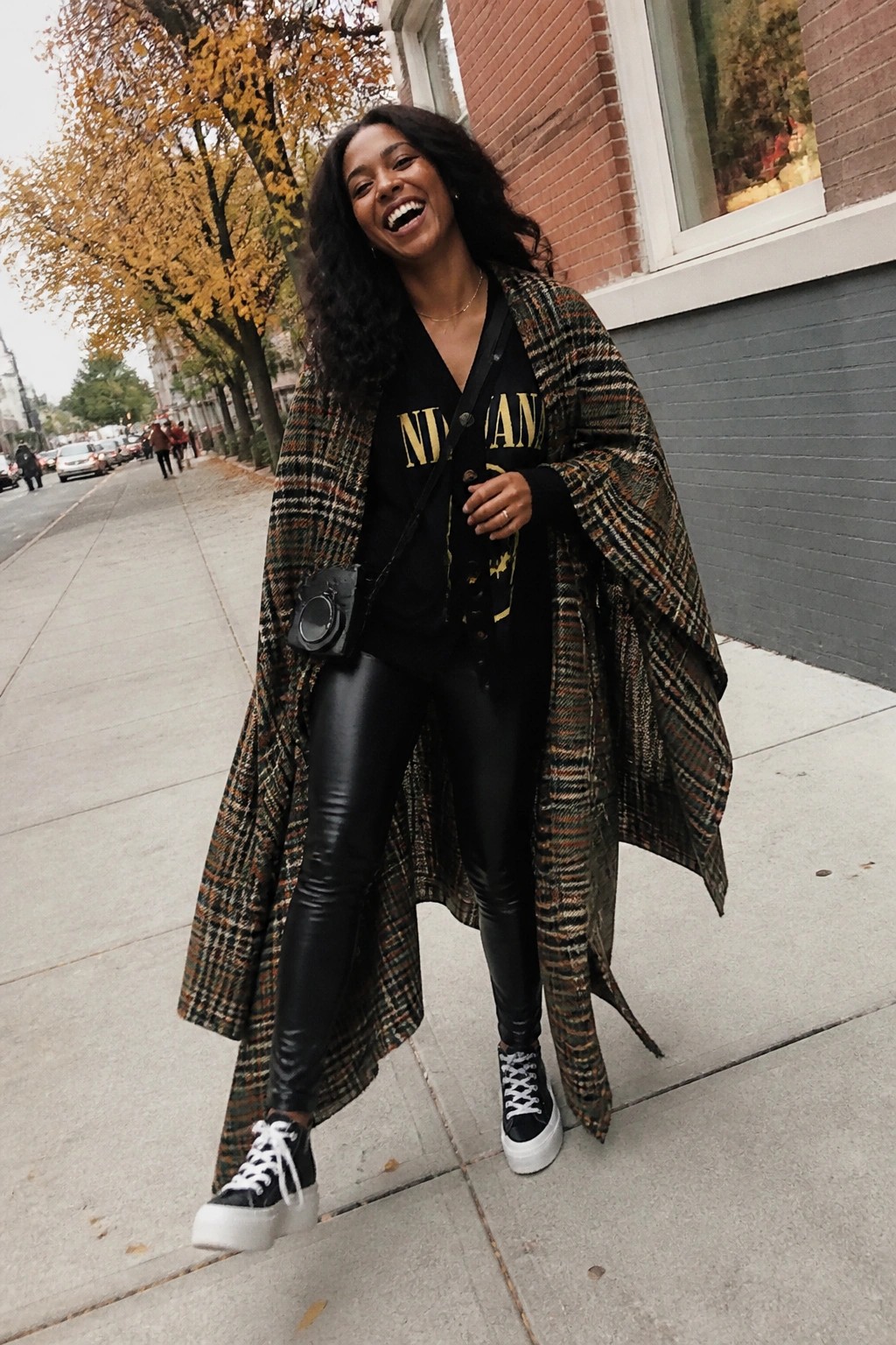 Woman wearing long plaid coat open over black Nirvana graphic tee and black leggings paired with platform sneakers and small crossbody bag.
