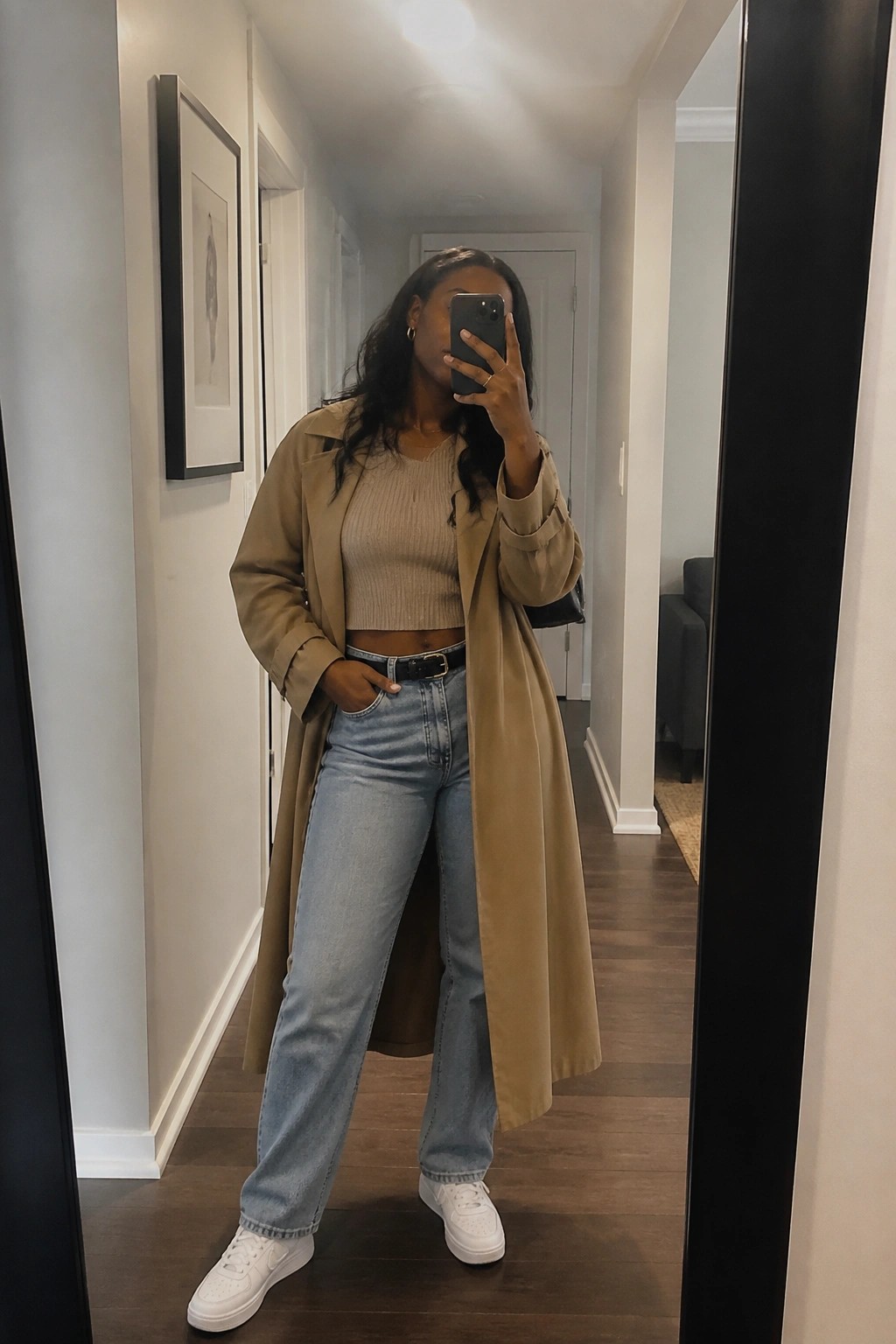 Woman in beige trench coat and jeans taking mirror selfie with phone
