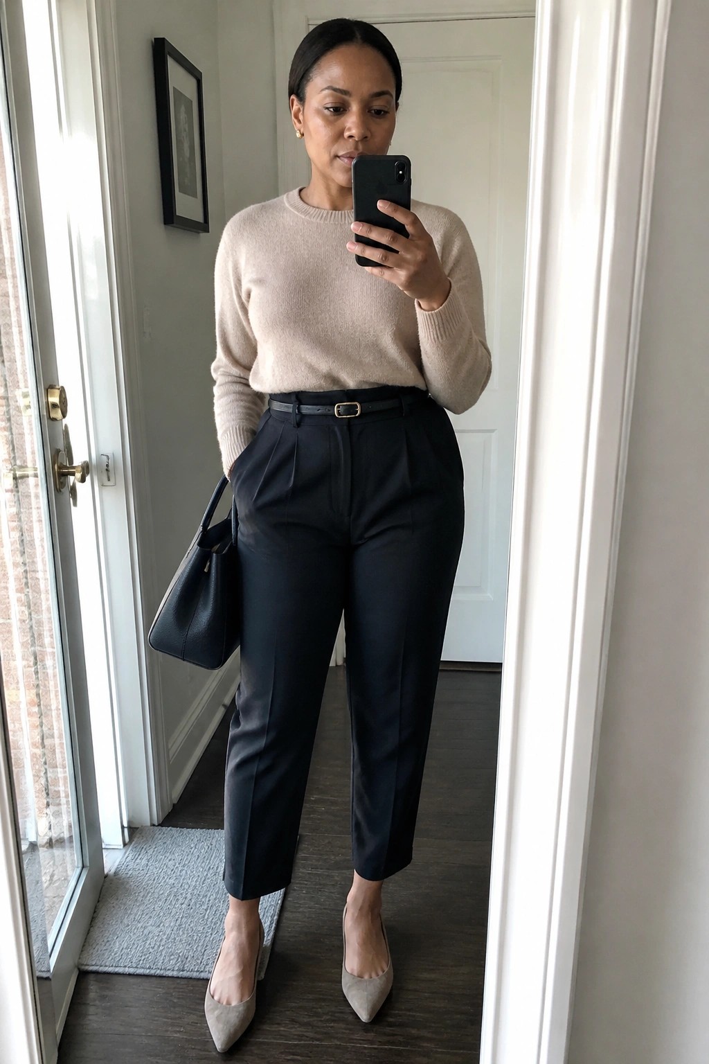 Woman in beige sweater and navy pants taking mirror selfie with handbag and heels