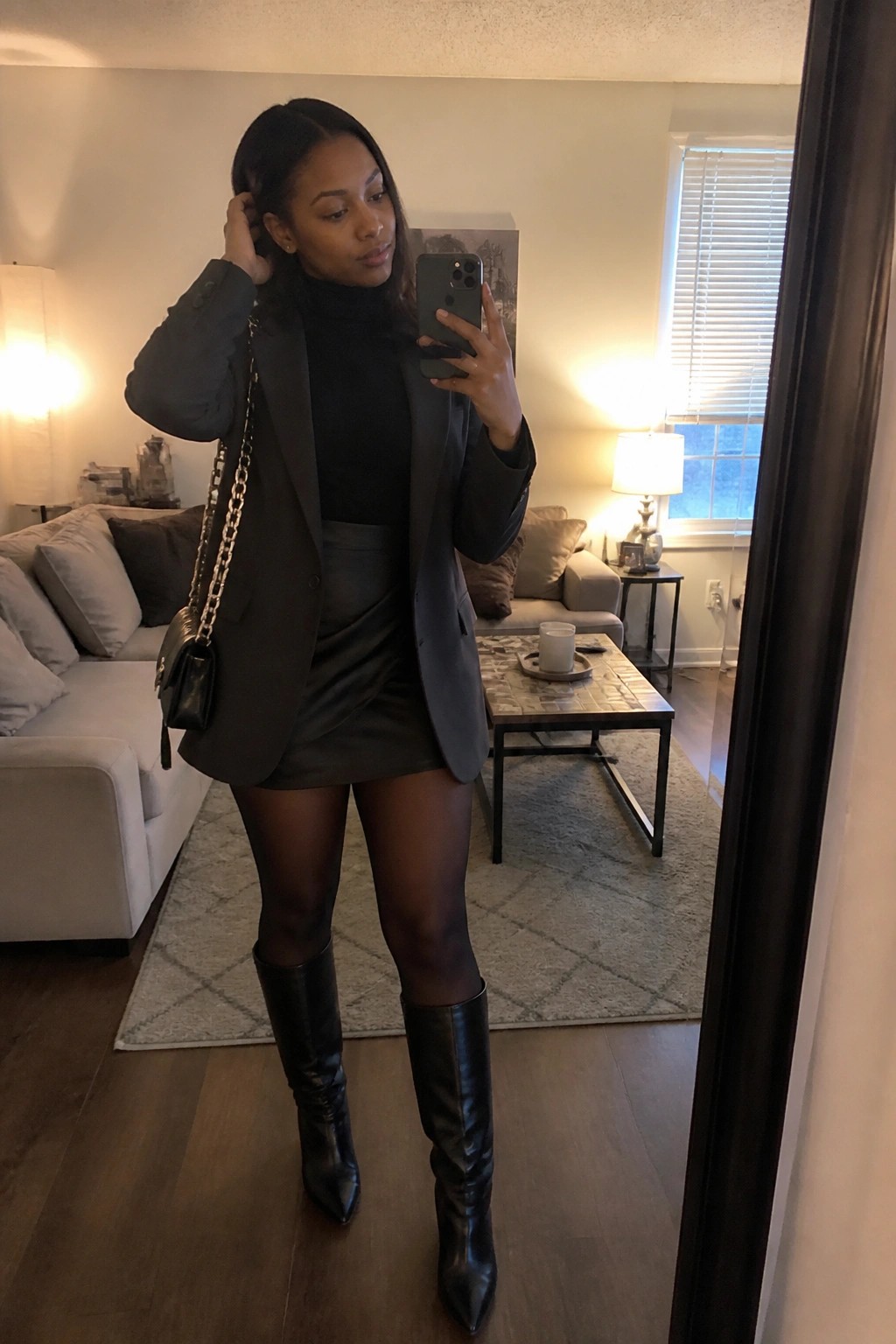 Woman in dark gray blazer over black turtleneck, gray mini skirt, black tights, and pointed black knee-high boots holding a phone for a mirror selfie with a black chain-strap bag.