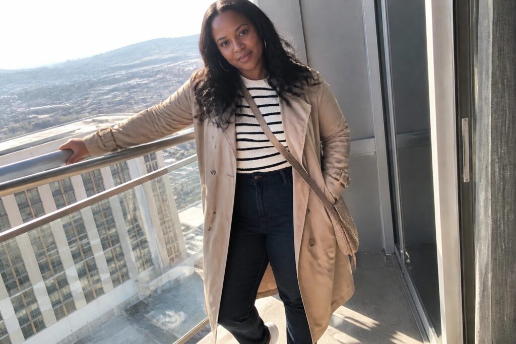 Woman in tan trench coat and jeans on balcony overlooking ocean at sunset