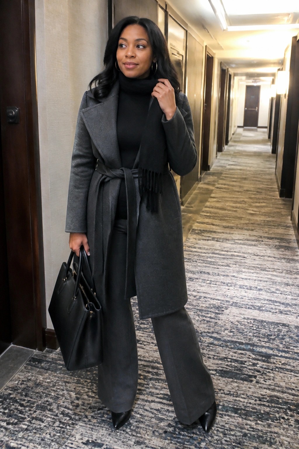 Woman in gray belted coat with black scarf holding handbag in hallway