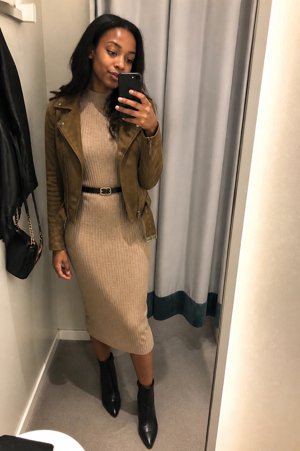 Woman in beige ribbed dress and brown jacket taking mirror selfie in fitting room.