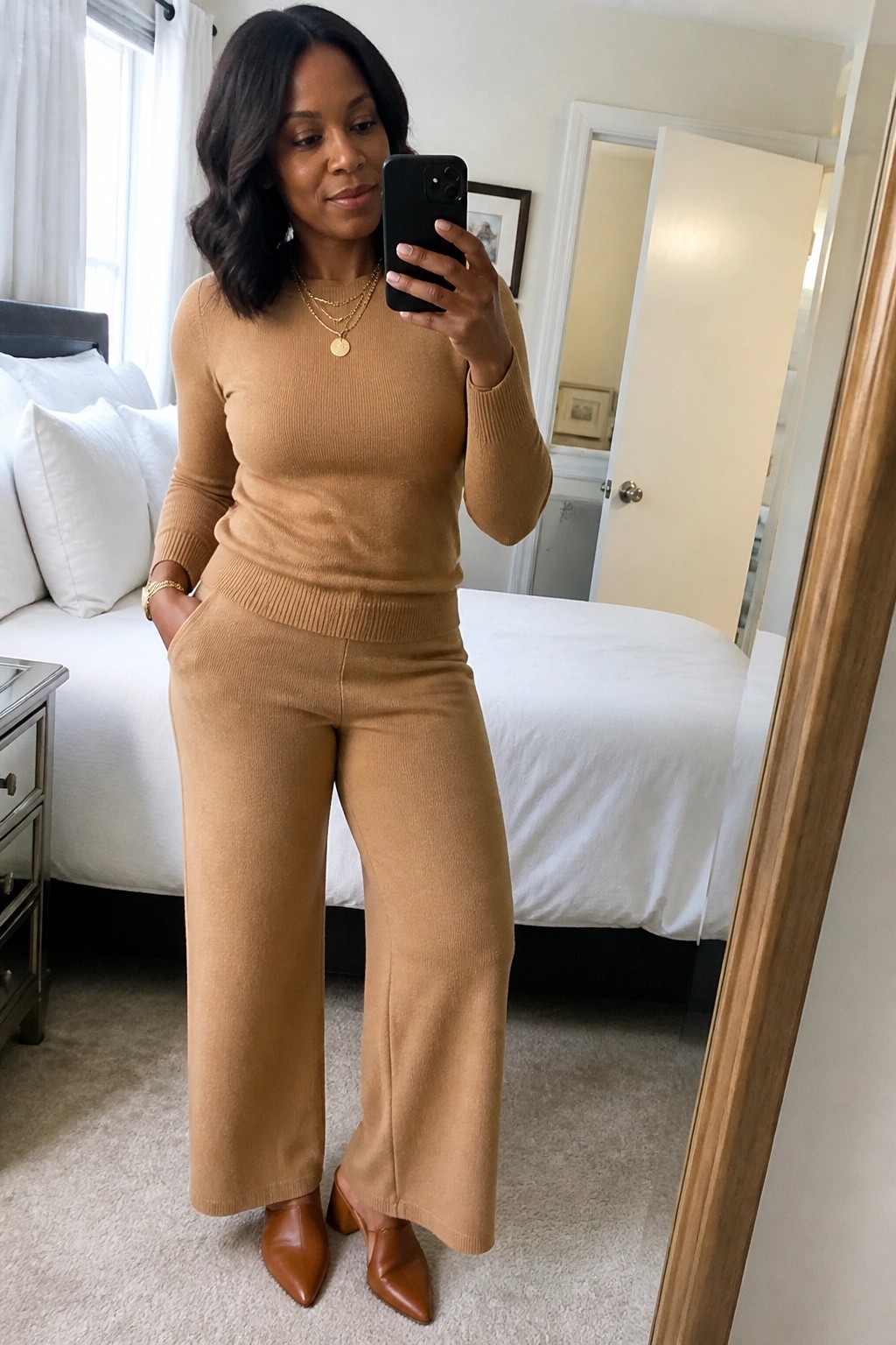 Woman in tan knit sweater set taking mirror selfie in bedroom