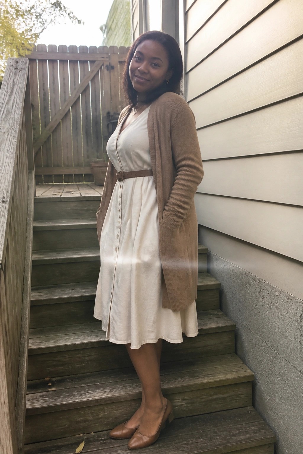 Woman in white dress and tan cardigan on wooden outdoor stairs by house.